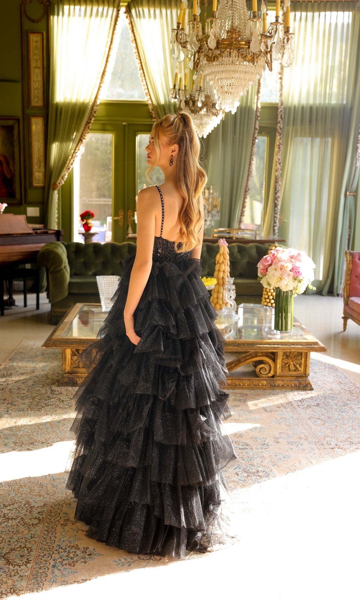 A woman wearing the Long Prom Dress 29232 by Ava Presley stands in a sunlit, elegant room with chandeliers, ornate furniture, and large windows draped in sheer curtains.